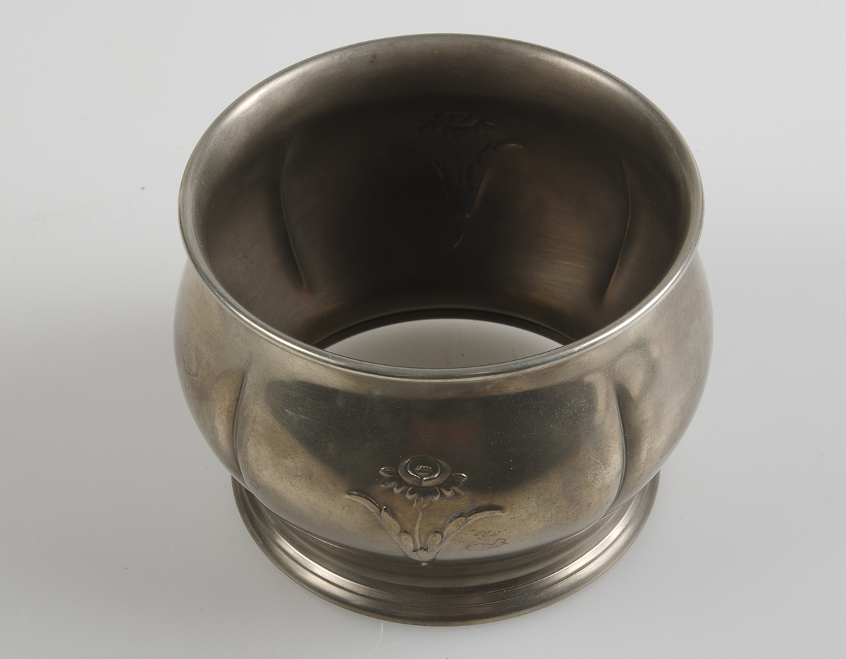 Del till sockerskål, rundel av gul metall med två blommor i relief. Botten och handtag saknas.
Sockerskål nodell nr 27 i katalog nr 20.