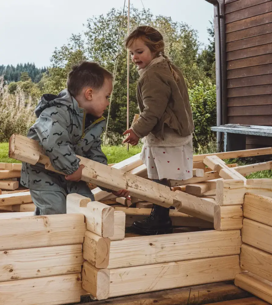 To barn står halveis inni et hus de bygger. Byggesett med laftestokker tilpasset deres størrelse. 