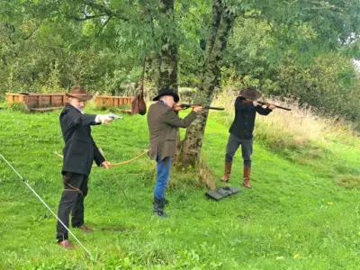 Tre menn kledd i westernklær og cowboyhatter sikter og skyter med rifler og pistol fra under et tre mot en blink utenfor bildet