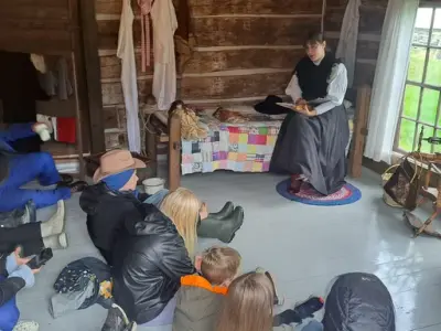 Barn sitter på gulvet i nybyggerstove et tømmerhus. På en seng med fargerikt lappeteppe sitter en kvinne i sort, langt skjørt, hvit bluse og trekantsjal og leser høyt og forteller fra en bok. 