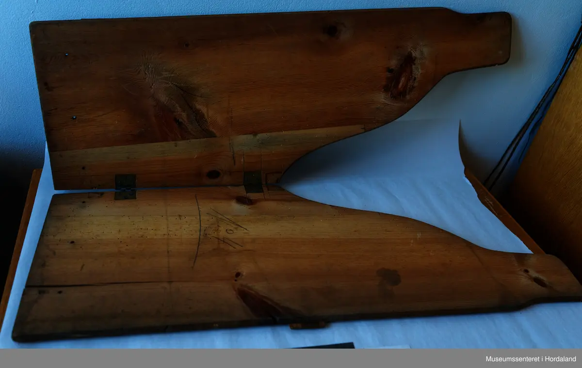 Bukseformer i tre. Kan brettes i to, Ble antakeligvis brukt til å tørke og forme ferdige underbukser før salg. Den har to hengsler.
Har to innfelte forsterkninger i tre, på tvers.