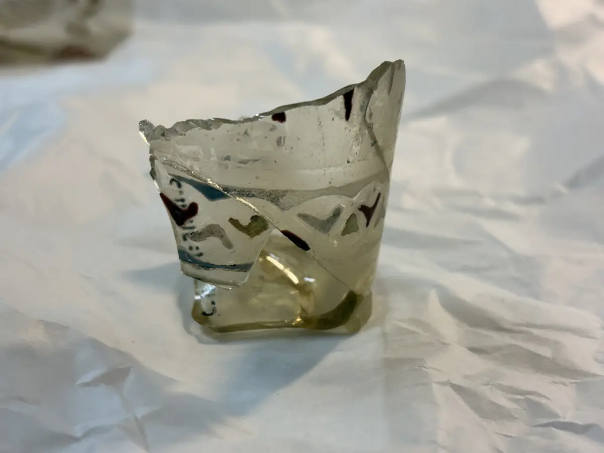 
Spår av röda, blå och vita färger, överst arabisk inskrift. 
Hela ytan är graverad med slingor, som troligtvis varit täckta med guld, och ur denna bakgrund framträder tre mänskliga figurer vars kläder varit dels emaljerade i rött och vitt, dels belagda med guld, som vid fyndet redan fallit bort. På glasets brätte finns tre vapensköldar som antyder att ägaren varit emir och mellan dessa en arabisk inskrift, som lytt: "Ära vare vår herre, sultanen, konungen , den rättvise, den vise, ära vare hans seger." Samma inskrift upprepas omedelbart under brättet. 

Nuvarande höjd 19, övre diam. C:a 11, nedre diam. C:a 6 cm. Inskrift översatt till svenska: "ära vare vår herre, sultanen, konungen, den rättvise, den vise, ära vare hans seger". 