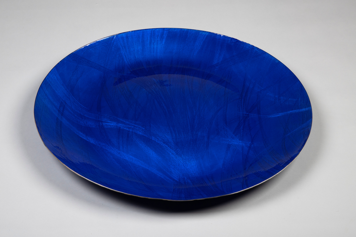 Fat i sølv med pålagt emalje. Sirkulær form med svakt hvelvet bunn og jevnt oppbuede sider. Over- og undersiden er dekket med blå emalje på frest bunn. Mønster i form av strålebunter som buer fritt over overflaten.