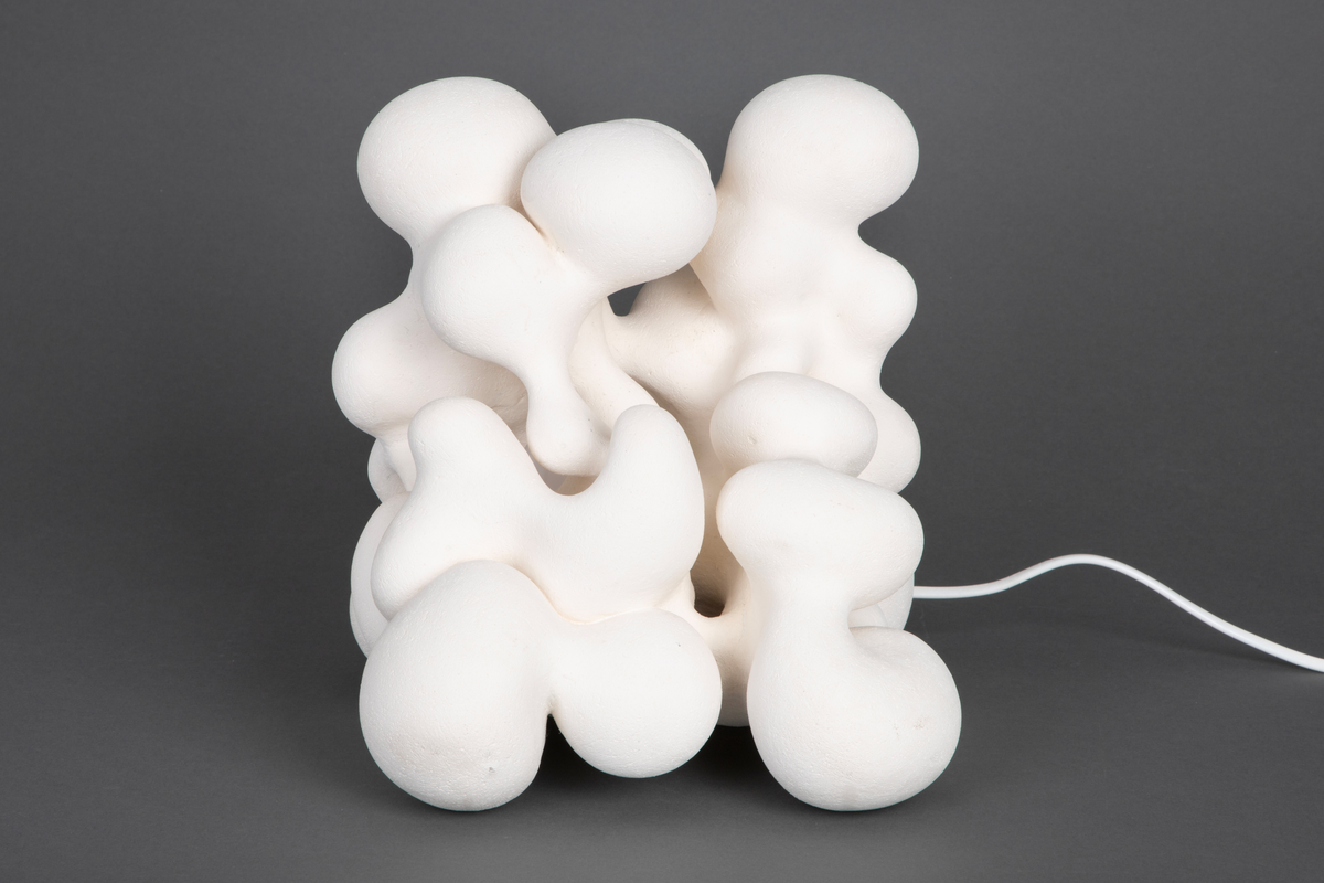 Hvit lampe i porselen. Formet av sammensmeltede bobleformer. Rund, gul lyspære inni, lyskilden er synlig fra utsiden. Skjult kabelfeste bak.