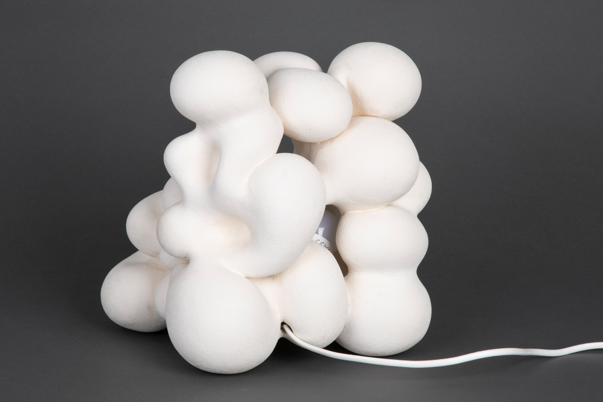 Hvit lampe i porselen. Formet av sammensmeltede bobleformer. Rund, gul lyspære inni, lyskilden er synlig fra utsiden. Skjult kabelfeste bak.