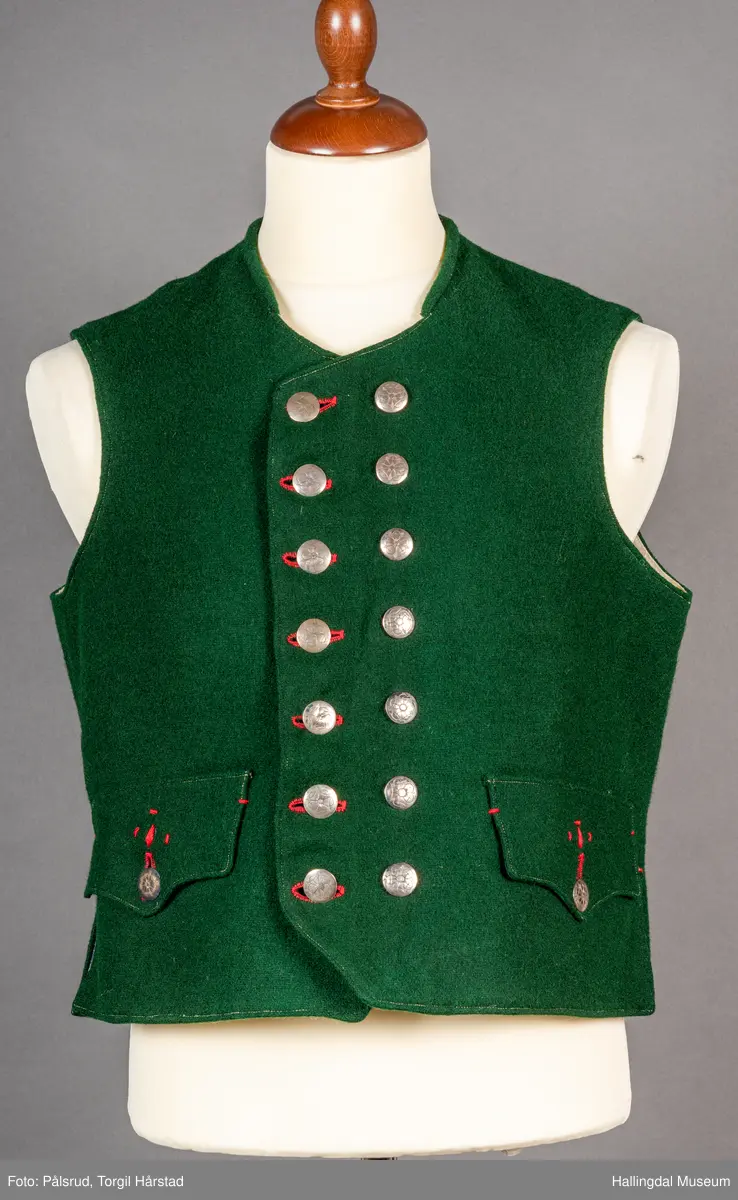 Vest til mann i grønn vadmel. Smal kanting i same stoff langs halsopningen. Dobbeltspent, 7 par knappar med fembladsrose festa med skinnlist. Knapphol sydd med raud ulltråd. 

Stikklomme med klaff på kvart forstykke. Lukking med knapp med fembladsrose, festa med skinnlist. På klaffen er knapphol med broderi i plattsaum over. Klaffen er forsterka med regels på kvar side. Knapphol, regels og prydsaum er sydd med raud ulltråd. 

Belegg langs framkant og nedkant på framstykke av kypervevd stoff. Sidesaum med splitt. Skuldersaum på skulder. Rygg i grønn vadmel. Saum midt bak, med raudt broderi over. 