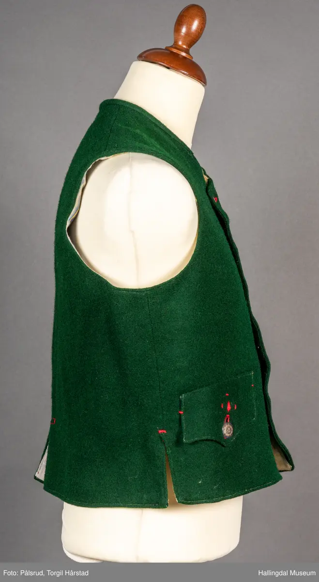 Vest til mann i grønn vadmel. Smal kanting i same stoff langs halsopningen. Dobbeltspent, 7 par knappar med fembladsrose festa med skinnlist. Knapphol sydd med raud ulltråd. 

Stikklomme med klaff på kvart forstykke. Lukking med knapp med fembladsrose, festa med skinnlist. På klaffen er knapphol med broderi i plattsaum over. Klaffen er forsterka med regels på kvar side. Knapphol, regels og prydsaum er sydd med raud ulltråd. 

Belegg langs framkant og nedkant på framstykke av kypervevd stoff. Sidesaum med splitt. Skuldersaum på skulder. Rygg i grønn vadmel. Saum midt bak, med raudt broderi over. 