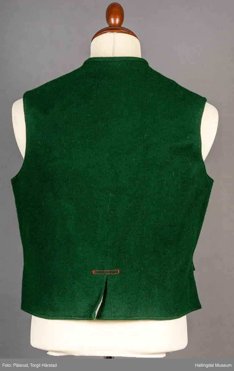 Vest til mann i grønn vadmel. Smal kanting i same stoff langs halsopningen. Dobbeltspent, 7 par knappar med fembladsrose festa med skinnlist. Knapphol sydd med raud ulltråd. 

Stikklomme med klaff på kvart forstykke. Lukking med knapp med fembladsrose, festa med skinnlist. På klaffen er knapphol med broderi i plattsaum over. Klaffen er forsterka med regels på kvar side. Knapphol, regels og prydsaum er sydd med raud ulltråd. 

Belegg langs framkant og nedkant på framstykke av kypervevd stoff. Sidesaum med splitt. Skuldersaum på skulder. Rygg i grønn vadmel. Saum midt bak, med raudt broderi over. 