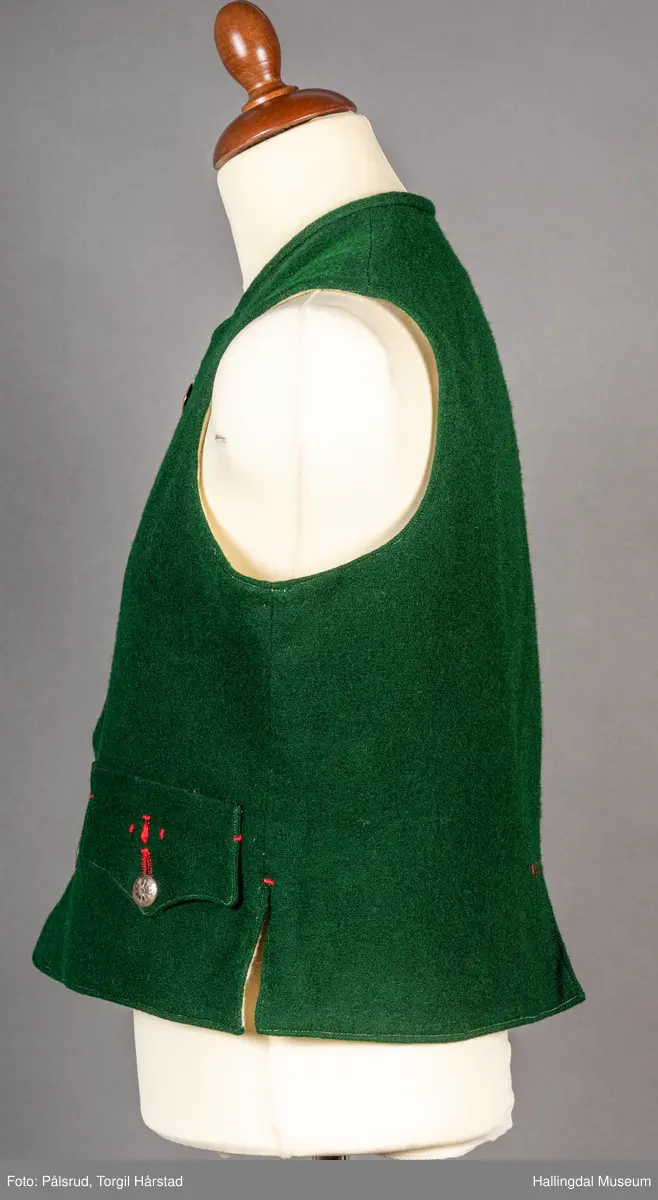 Vest til mann i grønn vadmel. Smal kanting i same stoff langs halsopningen. Dobbeltspent, 7 par knappar med fembladsrose festa med skinnlist. Knapphol sydd med raud ulltråd. 

Stikklomme med klaff på kvart forstykke. Lukking med knapp med fembladsrose, festa med skinnlist. På klaffen er knapphol med broderi i plattsaum over. Klaffen er forsterka med regels på kvar side. Knapphol, regels og prydsaum er sydd med raud ulltråd. 

Belegg langs framkant og nedkant på framstykke av kypervevd stoff. Sidesaum med splitt. Skuldersaum på skulder. Rygg i grønn vadmel. Saum midt bak, med raudt broderi over. 