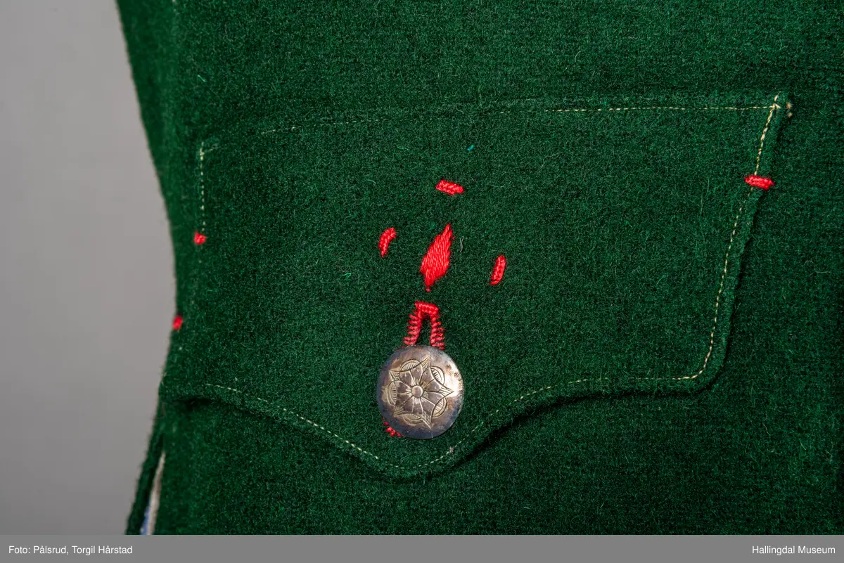 Vest til mann i grønn vadmel. Smal kanting i same stoff langs halsopningen. Dobbeltspent, 7 par knappar med fembladsrose festa med skinnlist. Knapphol sydd med raud ulltråd. 

Stikklomme med klaff på kvart forstykke. Lukking med knapp med fembladsrose, festa med skinnlist. På klaffen er knapphol med broderi i plattsaum over. Klaffen er forsterka med regels på kvar side. Knapphol, regels og prydsaum er sydd med raud ulltråd. 

Belegg langs framkant og nedkant på framstykke av kypervevd stoff. Sidesaum med splitt. Skuldersaum på skulder. Rygg i grønn vadmel. Saum midt bak, med raudt broderi over. 