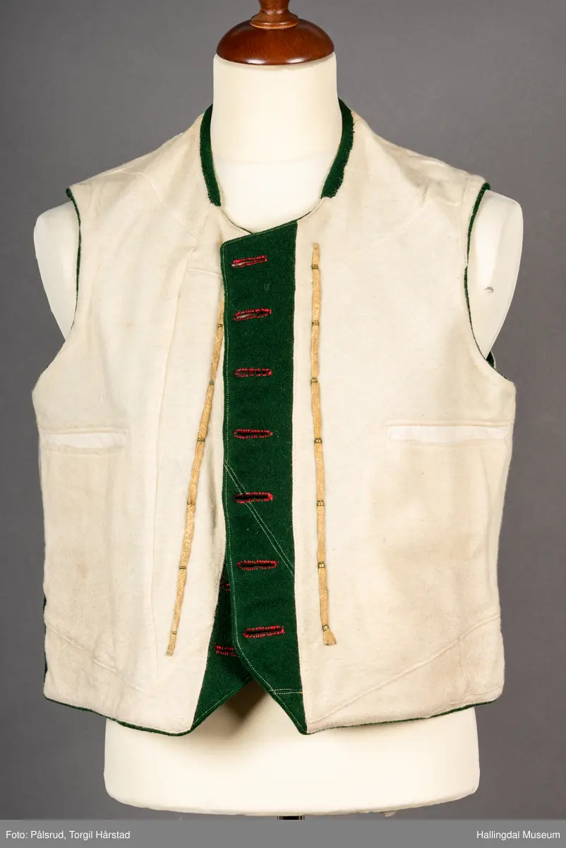 Vest til mann i grønn vadmel. Smal kanting i same stoff langs halsopningen. Dobbeltspent, 7 par knappar med fembladsrose festa med skinnlist. Knapphol sydd med raud ulltråd. 

Stikklomme med klaff på kvart forstykke. Lukking med knapp med fembladsrose, festa med skinnlist. På klaffen er knapphol med broderi i plattsaum over. Klaffen er forsterka med regels på kvar side. Knapphol, regels og prydsaum er sydd med raud ulltråd. 

Belegg langs framkant og nedkant på framstykke av kypervevd stoff. Sidesaum med splitt. Skuldersaum på skulder. Rygg i grønn vadmel. Saum midt bak, med raudt broderi over. 