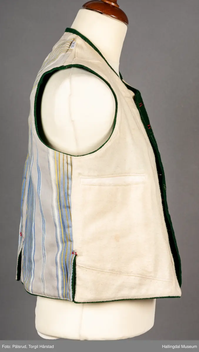 Vest til mann i grønn vadmel. Smal kanting i same stoff langs halsopningen. Dobbeltspent, 7 par knappar med fembladsrose festa med skinnlist. Knapphol sydd med raud ulltråd. 

Stikklomme med klaff på kvart forstykke. Lukking med knapp med fembladsrose, festa med skinnlist. På klaffen er knapphol med broderi i plattsaum over. Klaffen er forsterka med regels på kvar side. Knapphol, regels og prydsaum er sydd med raud ulltråd. 

Belegg langs framkant og nedkant på framstykke av kypervevd stoff. Sidesaum med splitt. Skuldersaum på skulder. Rygg i grønn vadmel. Saum midt bak, med raudt broderi over. 