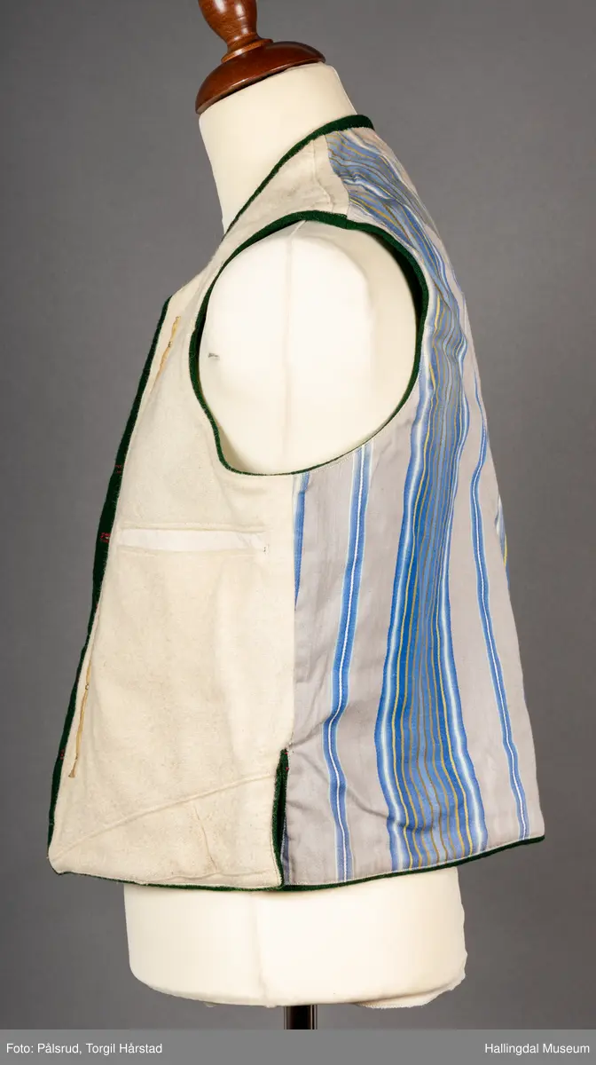 Vest til mann i grønn vadmel. Smal kanting i same stoff langs halsopningen. Dobbeltspent, 7 par knappar med fembladsrose festa med skinnlist. Knapphol sydd med raud ulltråd. 

Stikklomme med klaff på kvart forstykke. Lukking med knapp med fembladsrose, festa med skinnlist. På klaffen er knapphol med broderi i plattsaum over. Klaffen er forsterka med regels på kvar side. Knapphol, regels og prydsaum er sydd med raud ulltråd. 

Belegg langs framkant og nedkant på framstykke av kypervevd stoff. Sidesaum med splitt. Skuldersaum på skulder. Rygg i grønn vadmel. Saum midt bak, med raudt broderi over. 