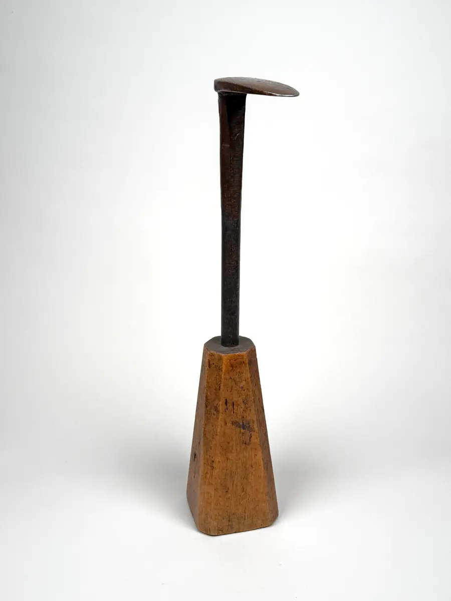 Skomakarverktyg av järn med lång hals och platt något ovalt huvud i vinkel mot halsen, på konisk fysidig fot av trä med avfasade sidor. 