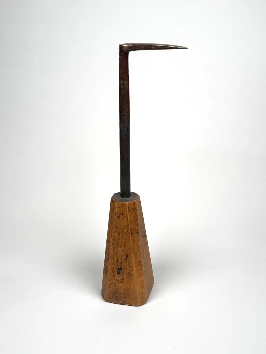 Skomakarverktyg av järn med lång hals och platt något ovalt huvud i vinkel mot halsen, på konisk fysidig fot av trä med avfasade sidor. 