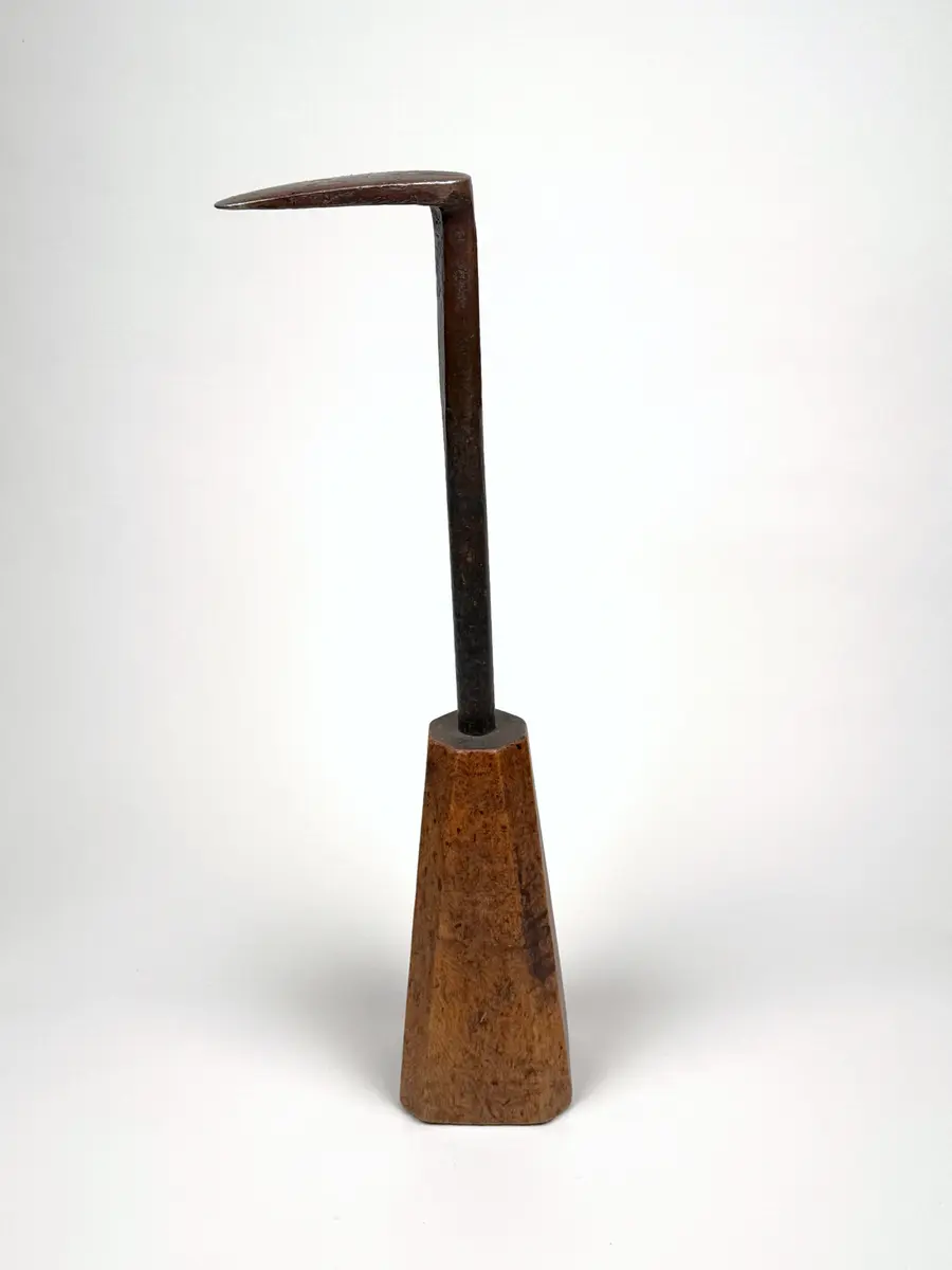 Skomakarverktyg av järn med lång hals och platt något ovalt huvud i vinkel mot halsen, på konisk fysidig fot av trä med avfasade sidor. 