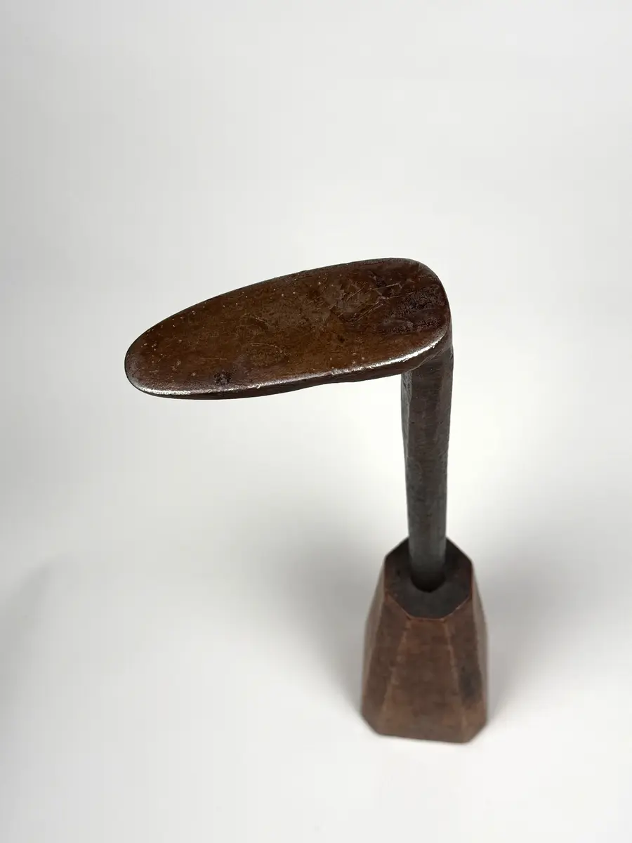 Skomakarverktyg av järn med lång hals och platt något ovalt huvud i vinkel mot halsen, på konisk fysidig fot av trä med avfasade sidor. 