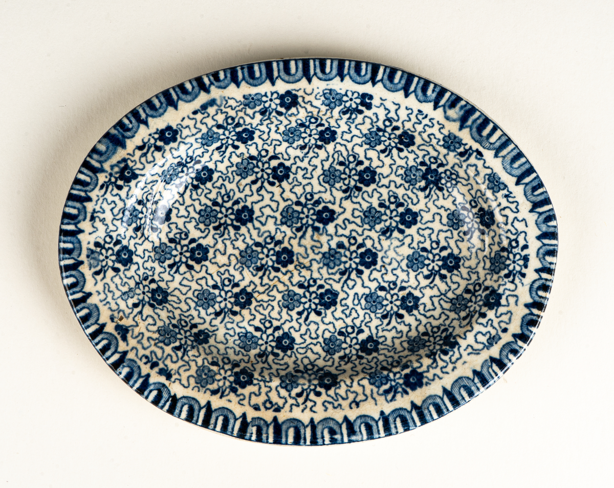 Fat, till dockservis. Ev flintgods. Glaserad. Vit med foralt mönster i blått. Ovalt. Vit och blå etikett.