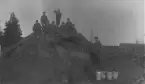 En grupp om närmare tjugo män samlade ovanpå och intill en koniskt uppbyggd vedstapel, sannolikt en kolmila. Männen är uppställda för fotografering och bär mörka kostymer, arbetskläder, hattar och kepsar typiska för sekelskiftet 1900. Några sitter högst upp på milan medan andra står längs sidorna och vid marken. I förgrunden syns fyra metallhinkar, möjligen avsedda för vatten i samband med kolningen. I bakgrunden framträder en större byggnad samt en kyrkspira, vilket antyder att platsen kan ha legat i anslutning till en bruksmiljö eller by. Bilden förefaller dokumentera ett arbetslag i samband med träkolstillverkning på landsbygden.