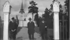 Svartvitt fotografi föreställande tre män stående vid en öppen grind. Männen är placerade mellan två vitmålade grindstolpar med dekorativa överstycken. De är klädda i mörka kostymer med slips och bär hattar.

Bakom männen leder en grusgång upp mot en kyrkobyggnad. Kyrkan har ett torn med spetsig spira, rundbågiga ljudöppningar i tornets övre del samt ett långhus med sadeltak. Byggnaden är uppförd i trä och omges av gräsytor och höga träd.