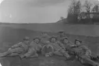 Grupporträtt av åtta uniformerade män som ligger och sitter tätt tillsammans på en gräsbevuxen mark i ett öppet landskap. De bär militäruniformer med mössor och flera av dem röker cigarett. Männen är avslappnat poserade och ser mot kameran. I bakgrunden syns öppna fält, en höjd med träd samt en byggnad, vilket tyder på att bilden är tagen utomhus i lantlig miljö. Fotografiet förefaller vara från tidigt 1900tal.