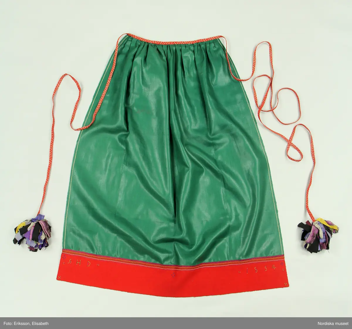 Huvudliggaren:
"Kvinnodräkt fr. Orsa sn, OfvanSiljans fgd, Dalarna.
a. Kjol med lif
b. Tröja         b. Sönderrifven å bakstycket.
c. Förkläde. Märkt: 'AHD 1854'
d. Öfverdel  [tecknat bomärke]
e. Halskläde [tecknat bomärke]
f. Strumpor
g. Skor
h. Band 5 st      h. valk, överklädd metalltråd
i. Hufvudkläde
j. Handskar
k. Bokkläde
l. Bjällernål. Af silver
m. Psalmbok. Tryckt i Göteb 1883."

Bilaga:
Finns ej.

Katalogkort:
Inga ytterligare uppgifter.

A. Livkjol,
Liv av högrött ylle med fågelmönster, importerat tyg trol engelskt av hög kvalitet, mitt fram kring öppningen  applikation av rött kläde med flerfärgat silkebroderi.4 par snörmaljor av tenn. Snörband av ljusbrunt skinn. Kjol av svartbrunt halvylle.
B. Tröja av svart kläde, lång modell. Sömmar sydda på maskin, stickningar runt kanterna för hand, nederkant och ärmuppslag har rå avklippt kant utan fåll.
Anm. Tröjan är helt ny och oanvänd, troligen uppsydd på beställning för förvärvet till museet för att få ihop en hel dräkt. Inte heller  psalmboken som ingår är gammal, den har tryckår 1883. Säljaren var en av Hazelius s.k. skaffare. Det var inte ovanligt att delar plockades ihop på bygden för att få ihop till en hel dräkt för museets räkning. 
Anm. I huvudliggaren står att litt b. är sönderriven  i ryggen, vilket inte stämmer.
/Berit Eldvik 2010-11-04

C. Förkläde "Glansmagd"
Av grön rask, nedtill en röd klädeslist som är broderad med silke i flera färger samt märkt AHD 1854.
D. Överdel. Smal utdragssöm över axeln med lila tråd markerar var ärmen börjar, ärmen avslutas med knäppta rynkor mot ärmlinning med nuggor i kanten och ett påsytt ca 1 cm brett mönstervävt band i blått och vitt. Enkelt halssprund utan knäppning.
/Berit Eldvik  2005-05

E: Halskläde i vit tuskaftad bomull . Parallella rynkningar med knäppning mot 5 cm hög rak krage som viks. Kragen prickar i langettsöm, bård i trädsöm, två rader efterstygn, nuggor utmed långsida,  Iträtt tuskaftsvävt knytband i rött och vitt, bred bård i gul, vit och rosa (fällningar) stopphålssöm, myrgångsbårder i kanterna. Dalknypplad spets fastsydd i framkant. Fållar maskinsydda för övrigt handsydd.
/Inga-Lill Eliasson 2007-08-01

K. Vitt kvadratiskt bokkläde i bomull, kypertdräll, rombiska former. Två stadkanter med gul tråd och två maskinsydda fållar.
/Inga-Lill Eliasson 2007-08-13