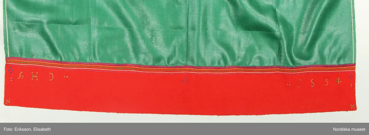 Huvudliggaren:
"Kvinnodräkt fr. Orsa sn, OfvanSiljans fgd, Dalarna.
a. Kjol med lif
b. Tröja         b. Sönderrifven å bakstycket.
c. Förkläde. Märkt: 'AHD 1854'
d. Öfverdel  [tecknat bomärke]
e. Halskläde [tecknat bomärke]
f. Strumpor
g. Skor
h. Band 5 st      h. valk, överklädd metalltråd
i. Hufvudkläde
j. Handskar
k. Bokkläde
l. Bjällernål. Af silver
m. Psalmbok. Tryckt i Göteb 1883."

Bilaga:
Finns ej.

Katalogkort:
Inga ytterligare uppgifter.

A. Livkjol,
Liv av högrött ylle med fågelmönster, importerat tyg trol engelskt av hög kvalitet, mitt fram kring öppningen  applikation av rött kläde med flerfärgat silkebroderi.4 par snörmaljor av tenn. Snörband av ljusbrunt skinn. Kjol av svartbrunt halvylle.
B. Tröja av svart kläde, lång modell. Sömmar sydda på maskin, stickningar runt kanterna för hand, nederkant och ärmuppslag har rå avklippt kant utan fåll.
Anm. Tröjan är helt ny och oanvänd, troligen uppsydd på beställning för förvärvet till museet för att få ihop en hel dräkt. Inte heller  psalmboken som ingår är gammal, den har tryckår 1883. Säljaren var en av Hazelius s.k. skaffare. Det var inte ovanligt att delar plockades ihop på bygden för att få ihop till en hel dräkt för museets räkning. 
Anm. I huvudliggaren står att litt b. är sönderriven  i ryggen, vilket inte stämmer.
/Berit Eldvik 2010-11-04

C. Förkläde "Glansmagd"
Av grön rask, nedtill en röd klädeslist som är broderad med silke i flera färger samt märkt AHD 1854.
D. Överdel. Smal utdragssöm över axeln med lila tråd markerar var ärmen börjar, ärmen avslutas med knäppta rynkor mot ärmlinning med nuggor i kanten och ett påsytt ca 1 cm brett mönstervävt band i blått och vitt. Enkelt halssprund utan knäppning.
/Berit Eldvik  2005-05

E: Halskläde i vit tuskaftad bomull . Parallella rynkningar med knäppning mot 5 cm hög rak krage som viks. Kragen prickar i langettsöm, bård i trädsöm, två rader efterstygn, nuggor utmed långsida,  Iträtt tuskaftsvävt knytband i rött och vitt, bred bård i gul, vit och rosa (fällningar) stopphålssöm, myrgångsbårder i kanterna. Dalknypplad spets fastsydd i framkant. Fållar maskinsydda för övrigt handsydd.
/Inga-Lill Eliasson 2007-08-01

K. Vitt kvadratiskt bokkläde i bomull, kypertdräll, rombiska former. Två stadkanter med gul tråd och två maskinsydda fållar.
/Inga-Lill Eliasson 2007-08-13