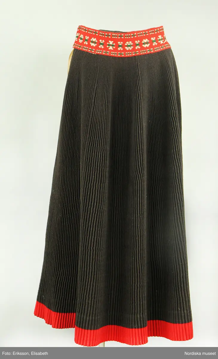 Huvudliggaren:
"Kvinnodräkt fr. Delsbo sn, Norra Helsinglands fgd, Helsingland.
a. "Ryntjeskjortel" af svart kläde med röd kant och bandväfd "kval" (linning). Kjolen "lurad"(goffrerad), skall förvaras lurad att ej vecken gå ut. 3 par fruntimmershänder hjälpas åt att "lura" den efter hvarje begagnande.
b. livstycke, rött med gröna ränder.
c. "Ryntjessärk" (rynksärk) af hvitt bomullstyg med "krus" (handbroderi) kring hals och händer.Begagnas sommartiden
d. "Bumsförkläde" af ylle, blårandigt med bandväf,"kval" i rödt och grönt.
e. "Förklesband" rödt och grönt ylle med "doppar" (tofsar) i ändarna. Lindas 1 à 2 hvarf kring lifvet och bindes framtill med stora nedhängande "locor" och ändar. Längd 262 cm, bredd 3,4 cm.
f. Kjolsäck med mässingslås och hake samt tennbroderi. Häktas under förklesbandet och hänger delvis under "försklet". Begagnas vanligen till förvaring af kyrkkryddor m.fl. kyrk-ellerkalassaker.
g. Halfhandskar af svart kläde med silkesbroderi. Begagnas sommartiden.
h. "Påshufva" hvardagsmössa af brokigt kattun. Begagnas med i nacken utslaget hår med några hårtestar hängande fritt framför öronen.
Till dräkten användes röda strumpor samt låga skor med höga klackar. Äfven kan användas silfersmycken kring halsen eller å  bröstet. En á två brokiga silkesdukar brukades omkring halsen nedstoppade under lifstycket, så att kruset kring halsen  på "ryntjessärken" sammanfästadt med ett löfspänne samt en smalare kant af hvarje sidenduk, blevo synliga. Vid handlederna sammanfästes ryntjessärken med röda sidenband eller länkknappar.

a. KJOL 
längd: 87 cm
midjeband bredd: 7 cm
av svart halvylle med svart linvarp och inslag av svart 1-tr. ullgarn, de 2 yttersta stadtrådarna är av oblekt lingarn. 4 goffrerade våder samt en slät framvåd som skarvats under förklädet med oblekt bomullslärft. 5 cm bred list av rött kläde i nederkanten runtom. Brett midjeband mönstervävt i rött och grönt ullgarn och vitt bomullsgarn. Knäppt vid framvådens vänstra sida med 2 hakar och hyskor av mässing.
b. LIVSTYCKE
längd: 39 cm
av halvylle i varptäckande kypert, varp av vitt lingarn, inslag av svart och grönt ullgarn med flaminslag av vitt, 2 fram-och sidstycken, 2 bakstycken avsmalnande till skörtveck med tre par tränsade förstärkningar av svart garn. I midjehöjd ett avsytt veck runtom, knäppt kant i kant med 8 par grova hakar och hyskor av mässing. Foder av ofärgad bomullslärft.
BE

c. ÖVERDEL
Längd: 39 cm. Längd: 58 cm - ärmlängd. Vidd: 56 cm - ärmvidd. Vidd: 114 cm. Höjd: 1,7 cm - kraghöjd. Material: Textil, bomull. Textil, linne - ärmlinning, krage. Bomullsgarn - broderi. Telnik: Tuskaft. Trådbundet broderi. Broderi, rätlinjig plattsöm. Handsömnad.
Överdel av tuskaftat vitt bomullstyg. Dubbelvikt bålstycke, isydda rektangulära halsspjäll. Täta breda stripade  rynkor mot halskrage i bomull, 1,7 cm hög. Kragen har broderier i rätlinjig plattsöm, vitt bomullsgarn över hela ytan, nuggor i kanten, langetterade bågar  i framkant. 16 cm långt sprund fram, tränsad förstärkning nedtill, 3 cm.  Vidsydd ärm med  infällda ärmspjäll, lagda veck över axeln fram och bak. Täta breda stripade rynkor mot 2,5 cm bred ärmlinning. Ärmlinningen har yttäckande vita broderier i rätlinjig plattsöm, rombiska former. Vita knytband i bomull i ärmlinningar och krage. Överdelen är handsydd.
/Inga-Lill Eliasson 2007-11-26


d. FÖRKLÄDE
längd=vådbredd: 82 cm
bredd: 97,5 cm
av halvylle, varp av svart lingarn, inslag i varptäckande kypert av svart 1-tr. ullgarn med smala ränder istarkt blått och en knappt synlig svart tråd med vita flamgarnsfläckar av bomull. Upptill stripade rynkor där de blå bårderna utnyttjats dekorativt i rynkningen. Mönstervävt midjeband i grönt på rött ullgarn , bandet knäpps med 2 hakar och hyskor i vänster sida.
I nederkantens båda hörn infodringar på insidan av svart bomullslärft, för att tyget inte skall rulla sig.



Berit Eldvik 2004