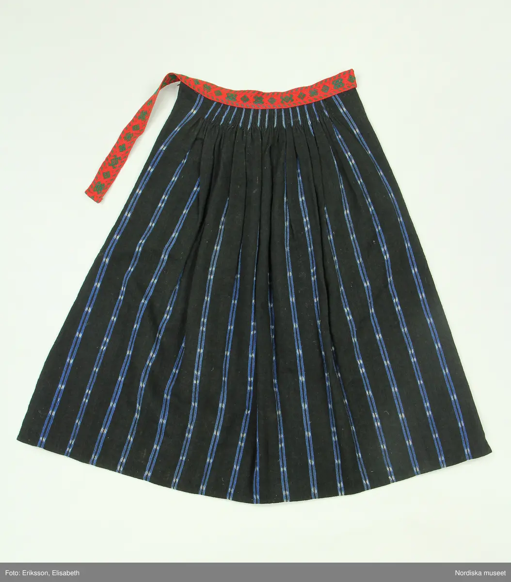 Huvudliggaren:
"Kvinnodräkt fr. Delsbo sn, Norra Helsinglands fgd, Helsingland.
a. "Ryntjeskjortel" af svart kläde med röd kant och bandväfd "kval" (linning). Kjolen "lurad"(goffrerad), skall förvaras lurad att ej vecken gå ut. 3 par fruntimmershänder hjälpas åt att "lura" den efter hvarje begagnande.
b. livstycke, rött med gröna ränder.
c. "Ryntjessärk" (rynksärk) af hvitt bomullstyg med "krus" (handbroderi) kring hals och händer.Begagnas sommartiden
d. "Bumsförkläde" af ylle, blårandigt med bandväf,"kval" i rödt och grönt.
e. "Förklesband" rödt och grönt ylle med "doppar" (tofsar) i ändarna. Lindas 1 à 2 hvarf kring lifvet och bindes framtill med stora nedhängande "locor" och ändar. Längd 262 cm, bredd 3,4 cm.
f. Kjolsäck med mässingslås och hake samt tennbroderi. Häktas under förklesbandet och hänger delvis under "försklet". Begagnas vanligen till förvaring af kyrkkryddor m.fl. kyrk-ellerkalassaker.
g. Halfhandskar af svart kläde med silkesbroderi. Begagnas sommartiden.
h. "Påshufva" hvardagsmössa af brokigt kattun. Begagnas med i nacken utslaget hår med några hårtestar hängande fritt framför öronen.
Till dräkten användes röda strumpor samt låga skor med höga klackar. Äfven kan användas silfersmycken kring halsen eller å  bröstet. En á två brokiga silkesdukar brukades omkring halsen nedstoppade under lifstycket, så att kruset kring halsen  på "ryntjessärken" sammanfästadt med ett löfspänne samt en smalare kant af hvarje sidenduk, blevo synliga. Vid handlederna sammanfästes ryntjessärken med röda sidenband eller länkknappar.

a. KJOL 
längd: 87 cm
midjeband bredd: 7 cm
av svart halvylle med svart linvarp och inslag av svart 1-tr. ullgarn, de 2 yttersta stadtrådarna är av oblekt lingarn. 4 goffrerade våder samt en slät framvåd som skarvats under förklädet med oblekt bomullslärft. 5 cm bred list av rött kläde i nederkanten runtom. Brett midjeband mönstervävt i rött och grönt ullgarn och vitt bomullsgarn. Knäppt vid framvådens vänstra sida med 2 hakar och hyskor av mässing.
b. LIVSTYCKE
längd: 39 cm
av halvylle i varptäckande kypert, varp av vitt lingarn, inslag av svart och grönt ullgarn med flaminslag av vitt, 2 fram-och sidstycken, 2 bakstycken avsmalnande till skörtveck med tre par tränsade förstärkningar av svart garn. I midjehöjd ett avsytt veck runtom, knäppt kant i kant med 8 par grova hakar och hyskor av mässing. Foder av ofärgad bomullslärft.
BE

c. ÖVERDEL
Längd: 39 cm. Längd: 58 cm - ärmlängd. Vidd: 56 cm - ärmvidd. Vidd: 114 cm. Höjd: 1,7 cm - kraghöjd. Material: Textil, bomull. Textil, linne - ärmlinning, krage. Bomullsgarn - broderi. Telnik: Tuskaft. Trådbundet broderi. Broderi, rätlinjig plattsöm. Handsömnad.
Överdel av tuskaftat vitt bomullstyg. Dubbelvikt bålstycke, isydda rektangulära halsspjäll. Täta breda stripade  rynkor mot halskrage i bomull, 1,7 cm hög. Kragen har broderier i rätlinjig plattsöm, vitt bomullsgarn över hela ytan, nuggor i kanten, langetterade bågar  i framkant. 16 cm långt sprund fram, tränsad förstärkning nedtill, 3 cm.  Vidsydd ärm med  infällda ärmspjäll, lagda veck över axeln fram och bak. Täta breda stripade rynkor mot 2,5 cm bred ärmlinning. Ärmlinningen har yttäckande vita broderier i rätlinjig plattsöm, rombiska former. Vita knytband i bomull i ärmlinningar och krage. Överdelen är handsydd.
/Inga-Lill Eliasson 2007-11-26


d. FÖRKLÄDE
längd=vådbredd: 82 cm
bredd: 97,5 cm
av halvylle, varp av svart lingarn, inslag i varptäckande kypert av svart 1-tr. ullgarn med smala ränder istarkt blått och en knappt synlig svart tråd med vita flamgarnsfläckar av bomull. Upptill stripade rynkor där de blå bårderna utnyttjats dekorativt i rynkningen. Mönstervävt midjeband i grönt på rött ullgarn , bandet knäpps med 2 hakar och hyskor i vänster sida.
I nederkantens båda hörn infodringar på insidan av svart bomullslärft, för att tyget inte skall rulla sig.



Berit Eldvik 2004