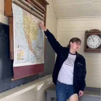Kvinne står og peiker på kart over USA. Kartet henger over en gammel skoletavle. På veggen bak henne en veggtavle 