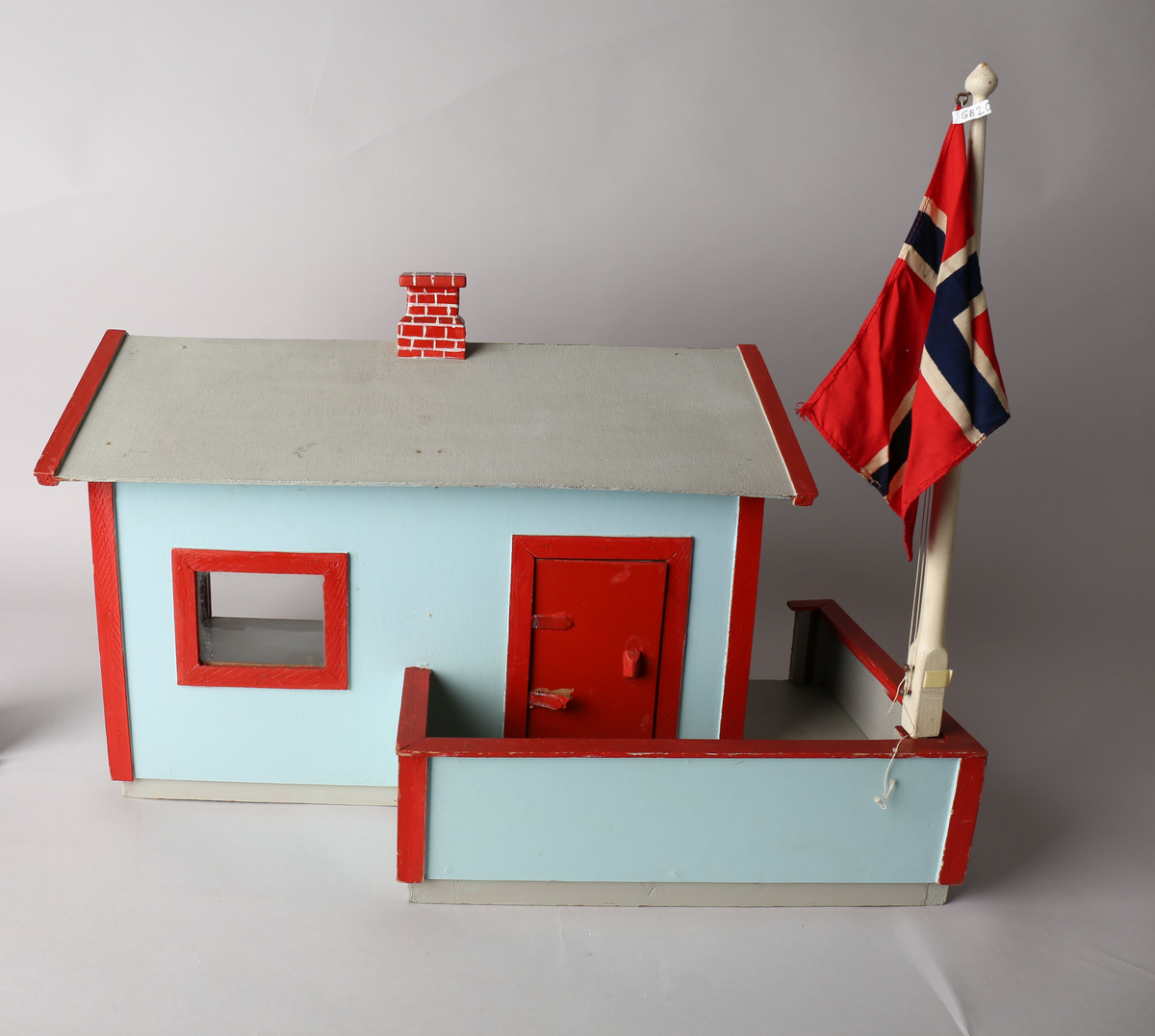 Rektangulært hus med påbygget veranda. Huset er malt lys blått med røde lister utvendig, innvendig tapet i brunbeige med hvite organiske striper og beide tapet med motiv av vegetasjon og blomster i hvitt, grønt og rødlige farger. Gulvet er malt grått og taket innvendig er umalt. Innvendige lister i trehvitt. Grått tak med rød pipe med påmalte hvite striper for å etterligne murstein. Sett forfra er dør ut til veranda i det venstre rommet. Innebygget kasse til flaggstang i venstre hjørne på veranda med avtagbar flaggstang. Norskt flagg heiset til topps i flaggstangen.
