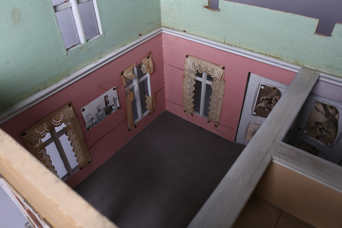 Rektangulært dukkehus i treverk med fire faste vegger. Huset består av fire rom og åpen loftsetasje. Totalt har huset ti vinduer og to doble dører som fungerer som enkeltdører. Utvendig er huset malt gult med hvite lister og brune dører og sprosser. Innvendig er gulvene malt grå, veggene rosa og gule med hvite innvendige sprosser. Gardiner i hvitt blondepapir og papirbilder er festet på veggene med tegnestifter. Veggene på loftet er malt mintgrønn.