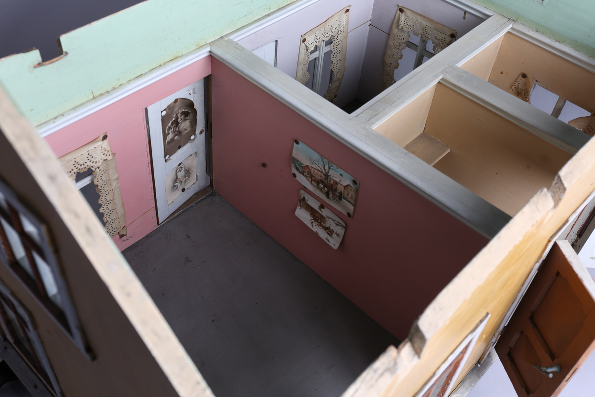 Rektangulært dukkehus i treverk med fire faste vegger. Huset består av fire rom og åpen loftsetasje. Totalt har huset ti vinduer og to doble dører som fungerer som enkeltdører. Utvendig er huset malt gult med hvite lister og brune dører og sprosser. Innvendig er gulvene malt grå, veggene rosa og gule med hvite innvendige sprosser. Gardiner i hvitt blondepapir og papirbilder er festet på veggene med tegnestifter. Veggene på loftet er malt mintgrønn.