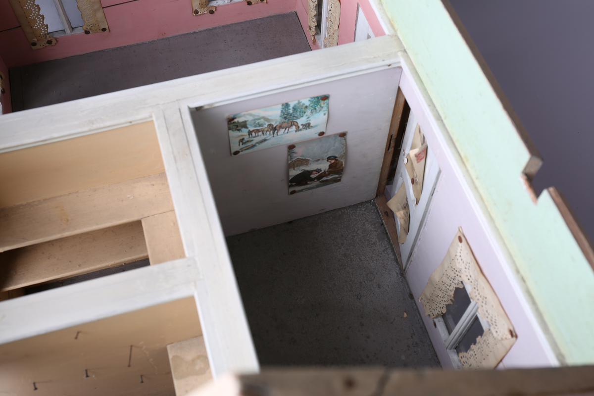 Rektangulært dukkehus i treverk med fire faste vegger. Huset består av fire rom og åpen loftsetasje. Totalt har huset ti vinduer og to doble dører som fungerer som enkeltdører. Utvendig er huset malt gult med hvite lister og brune dører og sprosser. Innvendig er gulvene malt grå, veggene rosa og gule med hvite innvendige sprosser. Gardiner i hvitt blondepapir og papirbilder er festet på veggene med tegnestifter. Veggene på loftet er malt mintgrønn.