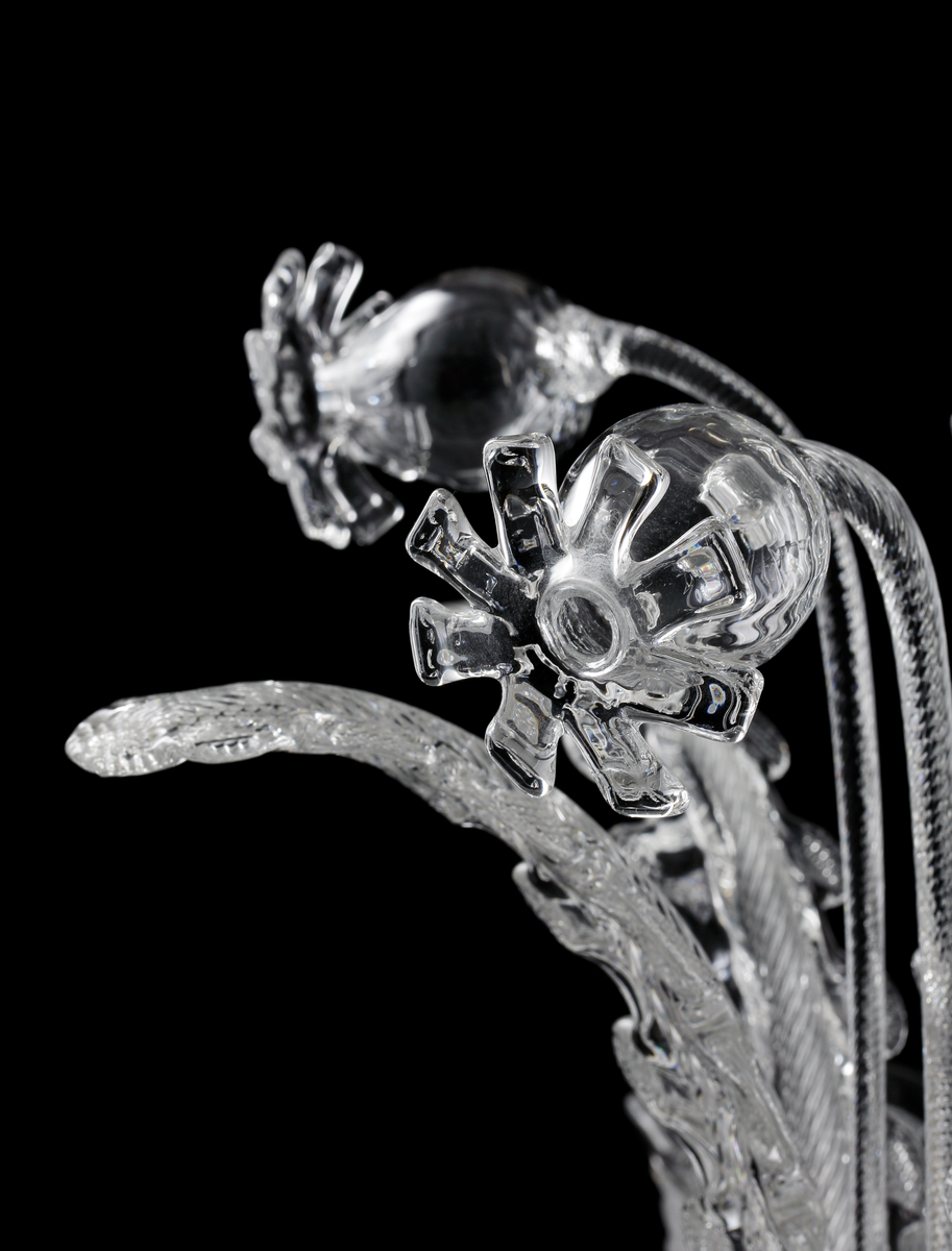 Takarmatur formgiven av Fritz Kurz. Underst en skål i pressat glas vari en infästningsanordning döljer sig. I denna sitter fem s-formade ljusarmar för elektriskt ljus placerade, tillsammans med sammanlagt tio stiliserade växt-dekorationer.