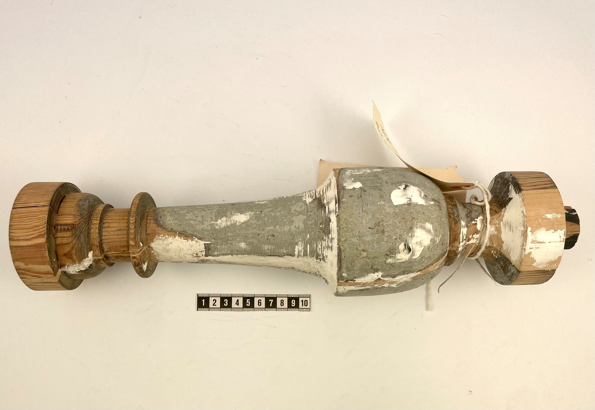 Baluster av ljust träslag i gott skick. Gråmålad med rester av vit färg över den grå färgen.
