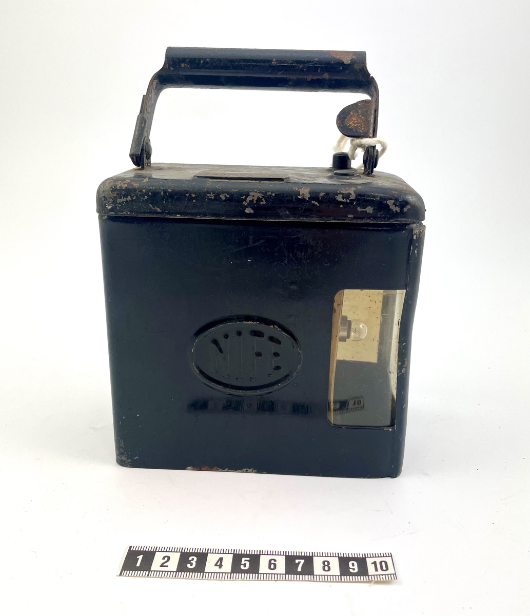 Svart elektrisk signalficklampa med handtag. Locket är löstagbart med en skruv på baksidan motsatt ljuset. Glödlampan täcks av glas. Handtaget är nedfällbart. Produktionsmärket NIFE står som ett emblem på båda sidor av lampan. På ovansidan står det "Arholma 1". I gott skick, något rostigt vid handtaget.