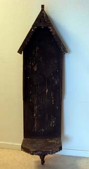 Bakstycke till en skulptur som tillsammans utgjort en kyrklig väggutsmyckning. Bakstycke av hopspikade brädor på vilken ett tak och ett fundament för en skulptur är fastspikat. Fundamentet i form av en hylla som sitter i fästad i bakstycket med hjälp av en snidad konsol. På kanten är utsågat ett dekorativt mönster. Taket i form av en takfot med list i vilken det är utsågat ett dekorativt mönster. Målad med brun målarfärg. I fundamentet spikar med vilka en nu försvunnen skulptur suttit fast.