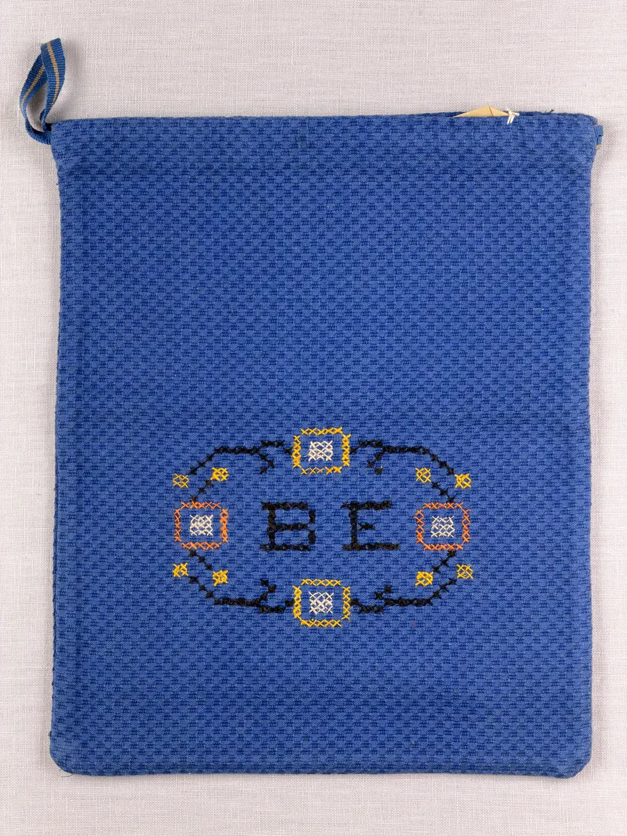 Tygpåse gjord av bomullsväv i sålldräll och broderi utfört med lingarn i korsstygn. Påsens öppning har en dragsko, 22mm bred dubbelvikt sydd med smygstygn, för förslutning med ett band. Påsen är hopsydd med vändsöm och raksöm.