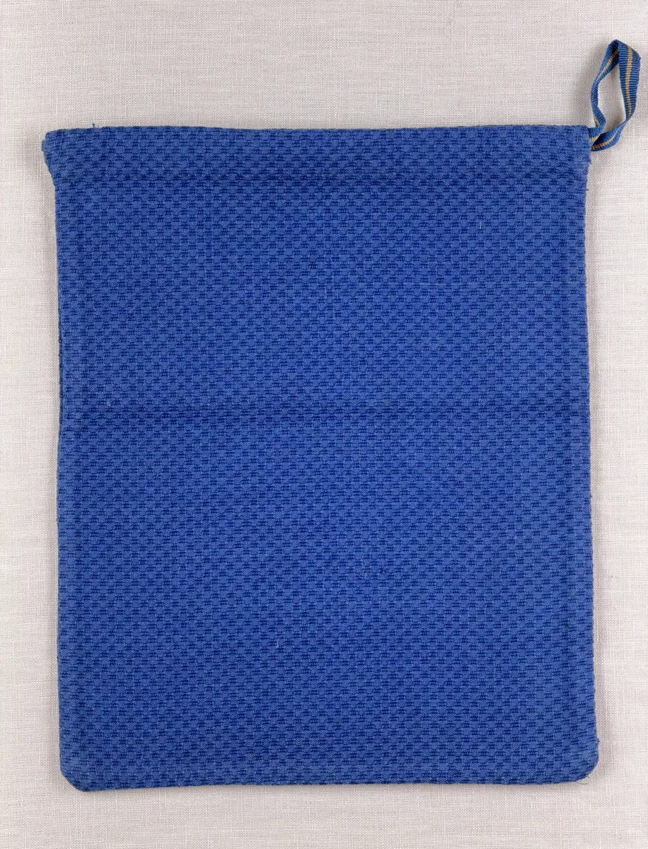 Tygpåse gjord av bomullsväv i sålldräll och broderi utfört med lingarn i korsstygn. Påsens öppning har en dragsko, 22mm bred dubbelvikt sydd med smygstygn, för förslutning med ett band. Påsen är hopsydd med vändsöm och raksöm.