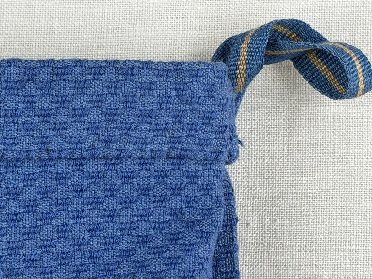 Tygpåse gjord av bomullsväv i sålldräll och broderi utfört med lingarn i korsstygn. Påsens öppning har en dragsko, 22mm bred dubbelvikt sydd med smygstygn, för förslutning med ett band. Påsen är hopsydd med vändsöm och raksöm.