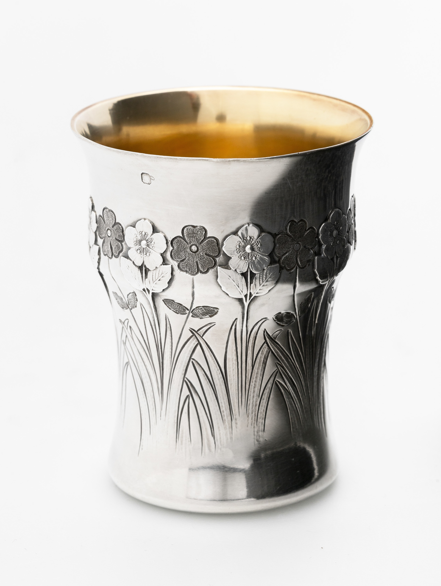 Konisk beger i sølv, dekorert med gravering og støpte detaljer. Svakt utbulet på øvre del, samt noe utfallende munningsrand. Yttersiden er dekorert med en frise av stilisert blomsterornamentikk samt bladverk. Blomstene er fremstilt vekselvis som lyse og mørke partier, vekslende utført i gravering og støping, slik at det oppstår et spill mellom positiv og negativ form. Innsiden er forgylt. 