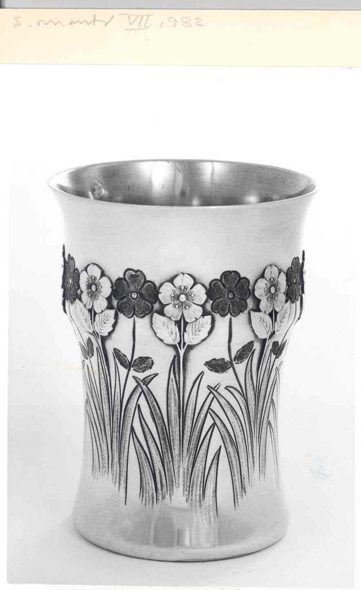 Konisk beger i sølv, dekorert med gravering og støpte detaljer. Svakt utbulet på øvre del, samt noe utfallende munningsrand. Yttersiden er dekorert med en frise av stilisert blomsterornamentikk samt bladverk. Blomstene er fremstilt vekselvis som lyse og mørke partier, vekslende utført i gravering og støping, slik at det oppstår et spill mellom positiv og negativ form. Innsiden er forgylt. 