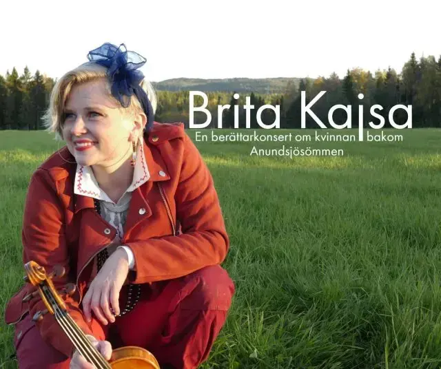 Kvinna med fiol sitter hukande på en äng med skog och berg i bakgrunden.