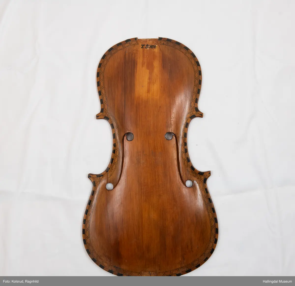 Lokk til hardingfele i gran. Spikka, uthola med fast basbjelke. Lakkert brun på oversida med svart staffelering rundt kanten. I følgje protokollen har den vore på ei hardingfele. 