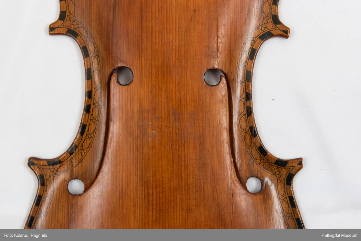 Lokk til hardingfele i gran. Spikka, uthola med fast basbjelke. Lakkert brun på oversida med svart staffelering rundt kanten. I følgje protokollen har den vore på ei hardingfele. 