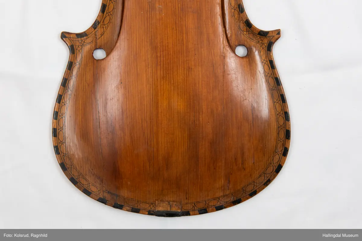 Lokk til hardingfele i gran. Spikka, uthola med fast basbjelke. Lakkert brun på oversida med svart staffelering rundt kanten. I følgje protokollen har den vore på ei hardingfele. 