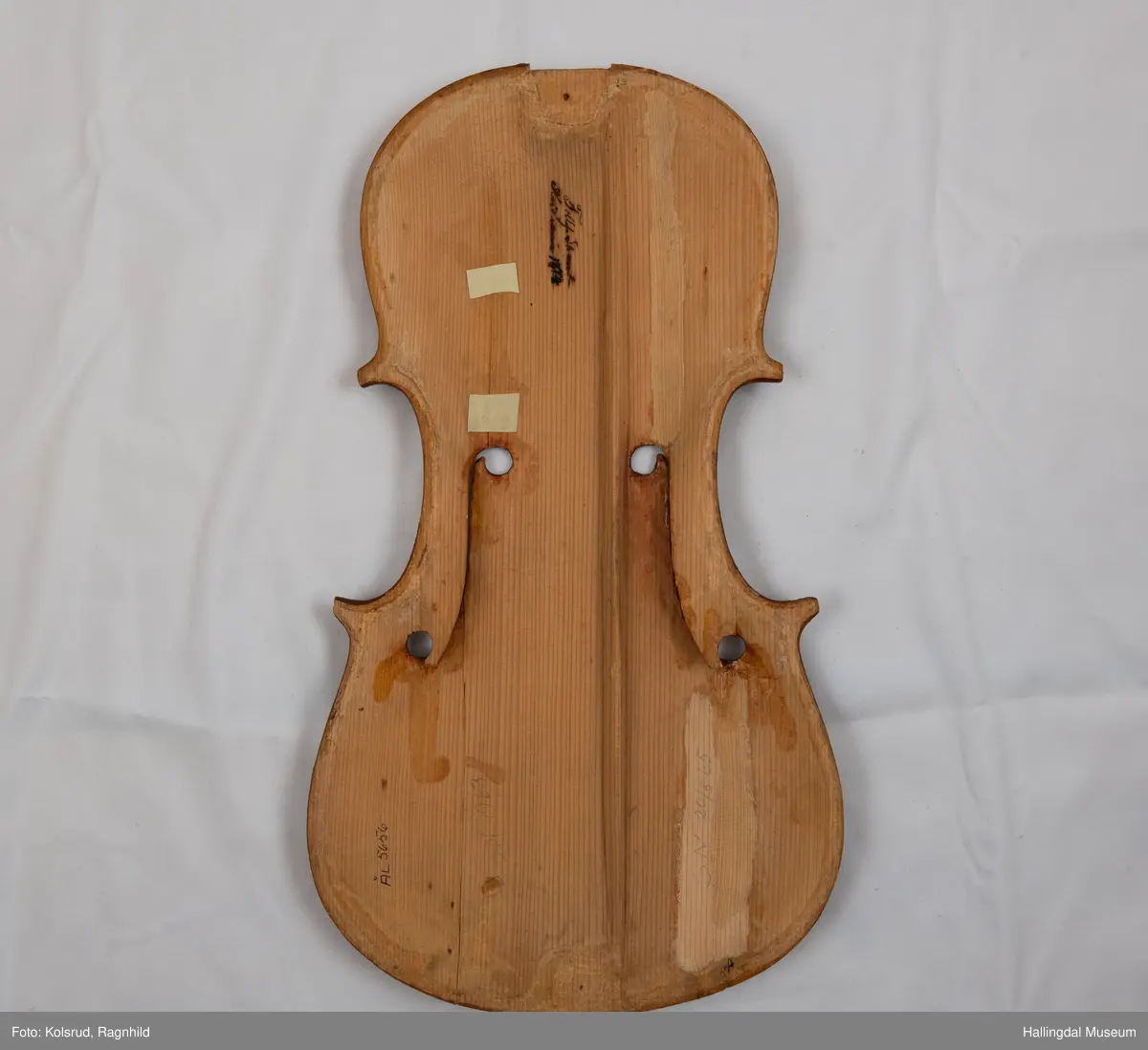 Lokk til hardingfele i gran. Spikka, uthola med fast basbjelke. Lakkert brun på oversida med svart staffelering rundt kanten. I følgje protokollen har den vore på ei hardingfele. 