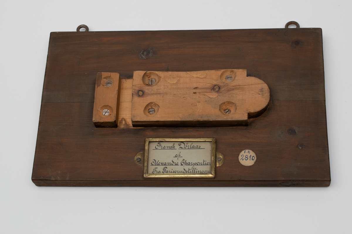 Rektangulært låsdeksel i støpt og fortinnet bronse. Horisontal grunnform bestående av låsekasse og sluttstykke. Forsiden viser en allegorisk fremstilling av en jente som synger, da som en personifisering av sangen.