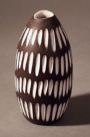 Vas  av lergods med avsmalnande mynning, dekorerad med fyra rader bestående av djupa paralella skåror med vit blank glasyr. Utsidan har för övrigt en svart halvmatt glasyr och insidan en vit blank glasyr, i botten stämplad 2131 UE Abg (=Ingrid Atterberg).